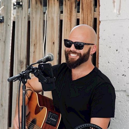 A person with a beard and sunglasses is playing an acoustic guitar and smiling, likely performing music outdoors.