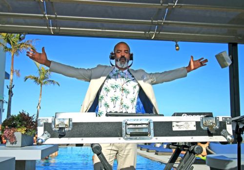 A person stands behind DJ equipment with arms outstretched, wearing headphones and a tropical shirt, with a pool and palm trees in the background.