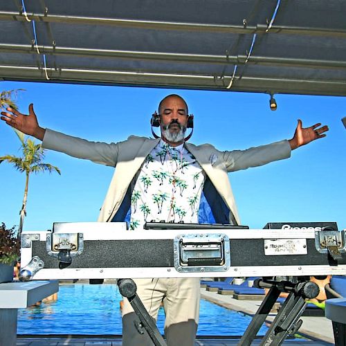 A person stands behind DJ equipment with arms outstretched, wearing headphones and a tropical shirt, with a pool and palm trees in the background.