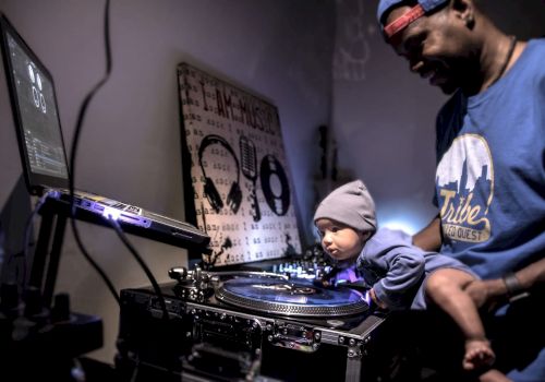 A man holds a baby who is interacting with DJ equipment; both are wearing blue hats and shirts.