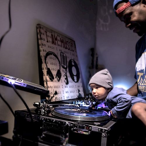 A man holds a baby who is interacting with DJ equipment; both are wearing blue hats and shirts.