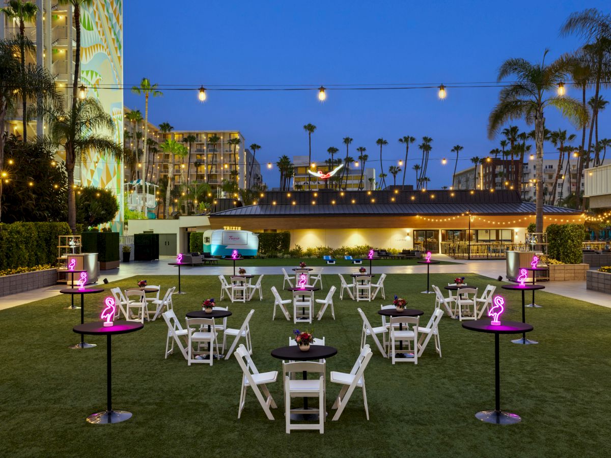 An outdoor event space is set up with round tables and chairs on a lawn, surrounded by string lights and palm trees, at dusk.