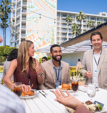 A group of people are having a conversation and eating at an outdoor table with drinks and sandwiches. The setting appears to be a sunny day.