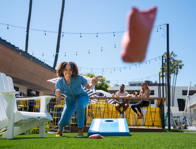 A person tosses a bean bag in a game of cornhole on a sunny outdoor patio, while others relax at a bar area in the background.