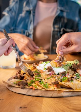 People are enjoying drinks and shared nachos at a table, with various hands reaching in to take some food.