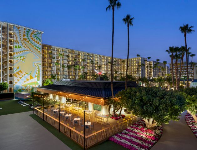 The image shows a nighttime view of a resort with a decorated tall building, an outdoor dining area, palm trees, and a well-maintained landscape.