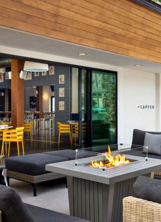 An outdoor patio with a fire pit, comfortable seating, and a view into an indoor dining area with yellow chairs and wall decorations.