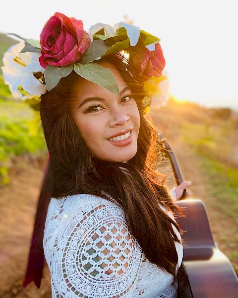 A woman with a flower crown and a guitar smiles outdoors, with sunlight illuminating the scene, suggesting an artistic or musical setting.