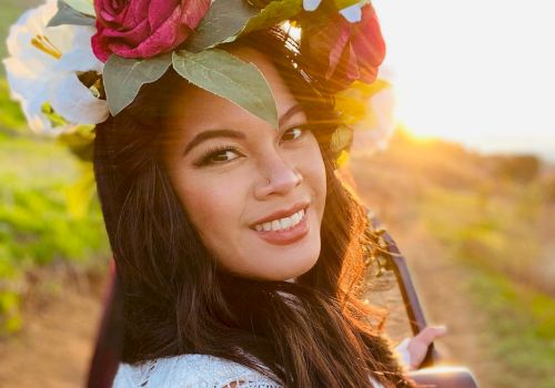 A woman with a flower crown and a guitar smiles outdoors, with sunlight illuminating the scene, suggesting an artistic or musical setting.