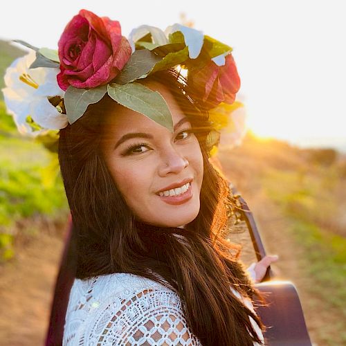 A woman with a flower crown and a guitar smiles outdoors, with sunlight illuminating the scene, suggesting an artistic or musical setting.