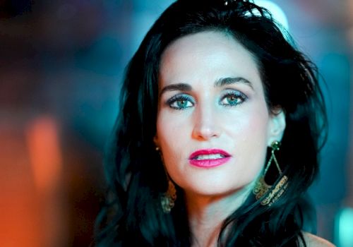 The image shows a woman with long dark hair, wearing gold earrings and makeup, with a blurred background that has blue and orange hues.