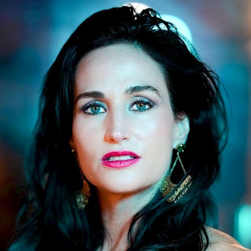 The image shows a woman with long dark hair, wearing gold earrings and makeup, with a blurred background that has blue and orange hues.