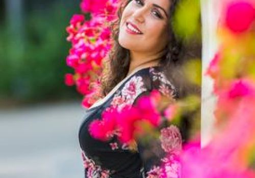 A woman, smiling, leans against a wall adorned with bright pink flowers, wearing a floral dress, with a blurred green background.