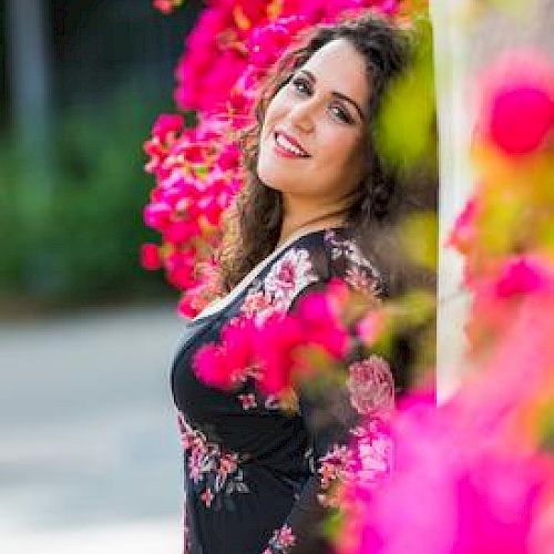 A woman, smiling, leans against a wall adorned with bright pink flowers, wearing a floral dress, with a blurred green background.