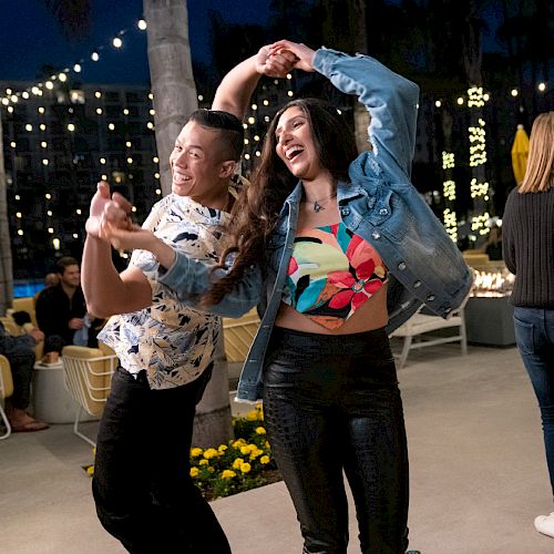 Two people are dancing joyfully with outdoor string lights and palm trees in the background. Others are seated nearby, enjoying the ambiance.