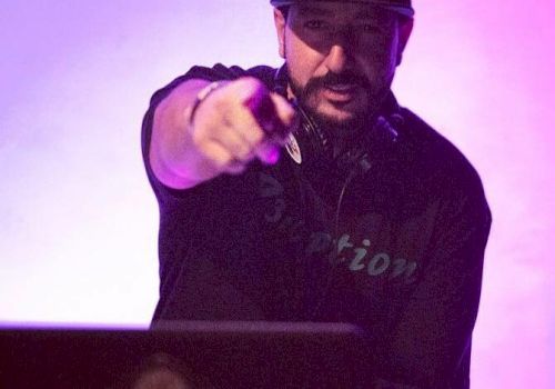 A DJ is performing on stage, wearing a hat and headset, pointing toward the camera, with a purple-lit background.