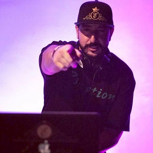 A DJ is performing on stage, wearing a hat and headset, pointing toward the camera, with a purple-lit background.