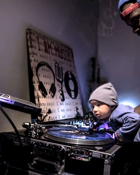 A man holding a baby is standing next to DJ equipment, with the baby touching the turntable.