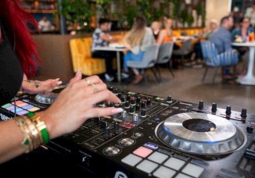 A DJ is playing music on a mixer, with people seated and chatting in the background at a lively venue.