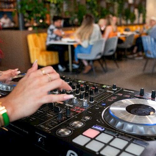 A DJ is playing music on a mixer, with people seated and chatting in the background at a lively venue.