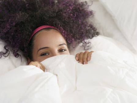 A person with curly hair and a headband is partially hidden under a white blanket, showing just their eyes and hands in a cozy setting.