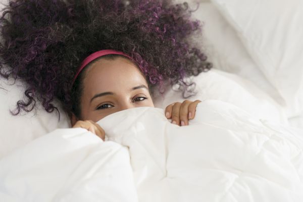 A person with curly hair and a headband is partially hidden under a white blanket, showing just their eyes and hands in a cozy setting.