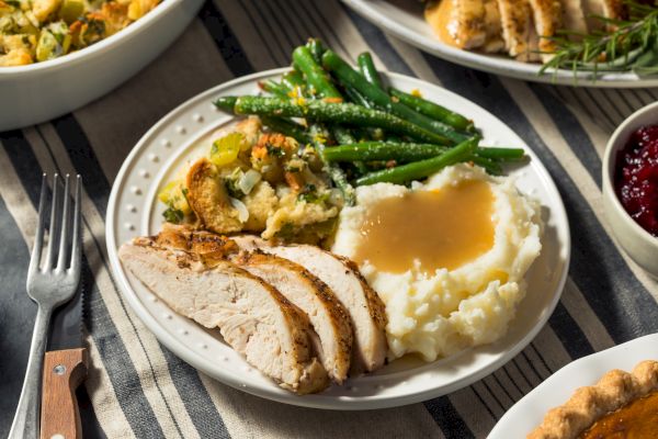 The image shows a plate with sliced turkey, mashed potatoes with gravy, green beans, and stuffing on a striped cloth.