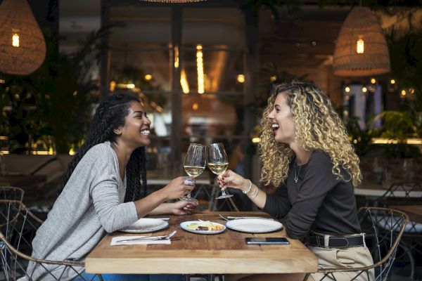 Two women at a restaurant, laughing and toasting with glasses of white wine, enjoying a meal together with plates of food on the table.