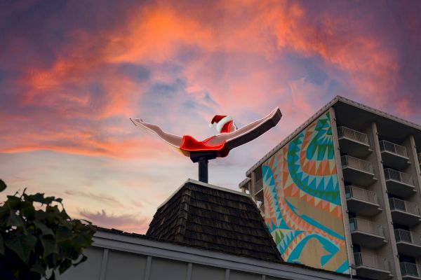 A vibrant sky at sunset behind a rooftop sculpture of a swimmer diving alongside a mural on a tall building.