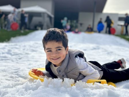 A child is laying on the snow with a sled, smiling, in front of a modern building. Other people are in the background.