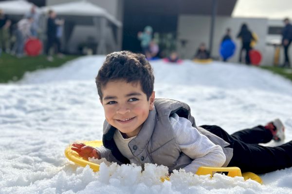 A child is laying on the snow with a sled, smiling, in front of a modern building. Other people are in the background.