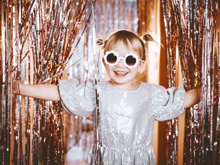 A child in a shiny silver dress and round glasses smiles, standing amidst sparkling, metallic streamers, creating a festive atmosphere.