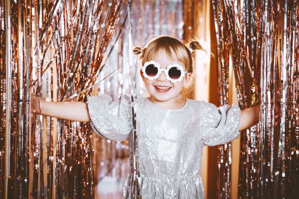 A child in a shiny silver dress and round glasses smiles, standing amidst sparkling, metallic streamers, creating a festive atmosphere.