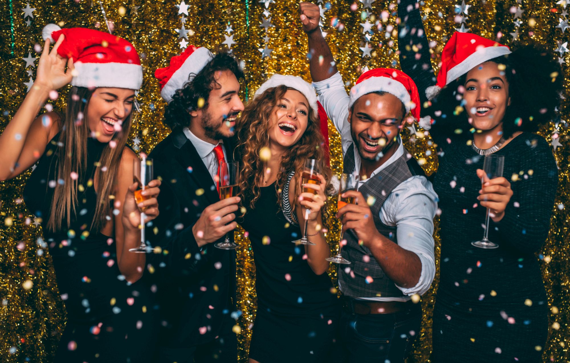 A group of five people wearing Santa hats, holding drinks, and celebrating with confetti against a festive, glittery backdrop.