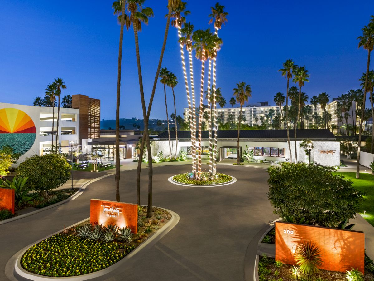 This image shows a beautifully landscaped hotel entrance with tall palm trees, modern art, and well-lit walkways during the evening.