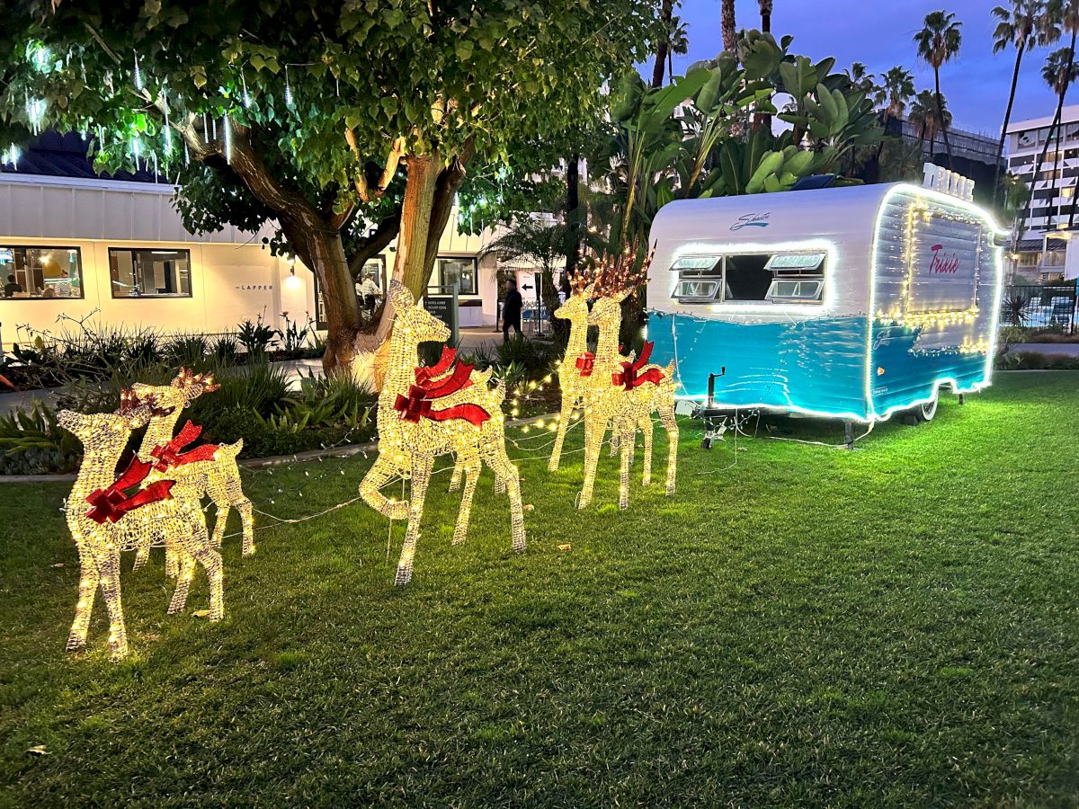 The image shows a festive holiday setup with illuminated reindeer decorations and a blue-and-white trailer on a grassy area, surrounded by buildings and trees.