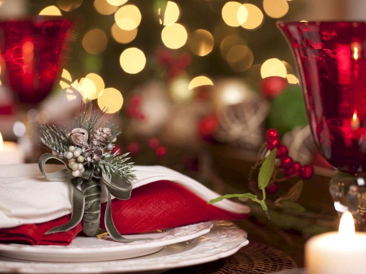 A festive table setting with red glassware, white plates, red napkins, holiday decorations, and candles, with blurry lights in the background.