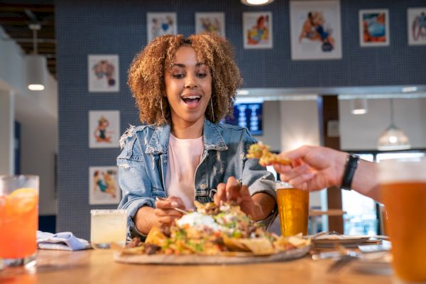 A person joyfully reaches for nachos at a restaurant, surrounded by drinks and colorful wall art in the background.