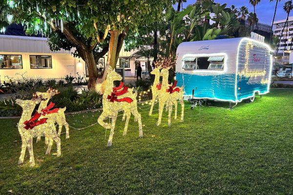 Lit-up reindeer decorations with red ribbons stand on grass beside a blue and white holiday trailer; trees and buildings are in the background.