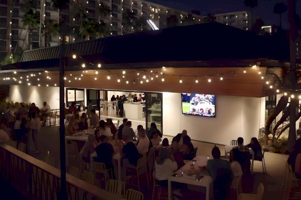 An outdoor restaurant scene at night with diners under string lights, a TV screen showing a sports game, and a modern building background.