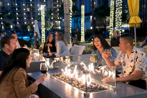 A group of people are enjoying drinks around a modern outdoor fireplace at night, with decorated trees and lights in the background.