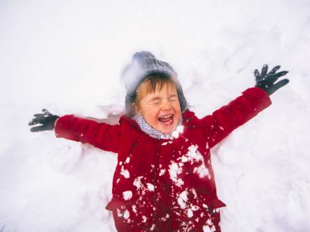 A child in a red coat joyfully makes a snow angel, wearing gloves and a hat, surrounded by snow.