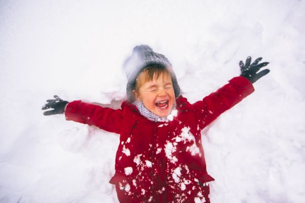 A child in a red coat joyfully makes a snow angel, wearing gloves and a hat, surrounded by snow.