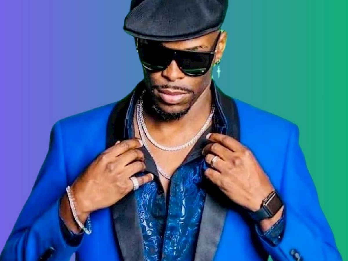 A person wearing a blue suit, black hat, sunglasses, and jewelry stands against a gradient background of purple and green shade, looking stylish.