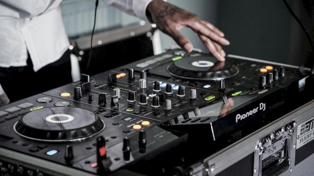 A DJ is mixing music on a Pioneer DJ controller with various knobs and buttons being adjusted. The DJ's hand is on one of the jog wheels.