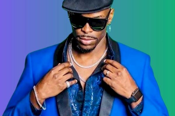 A person wearing a blue suit, hat, and sunglasses, accessorized with jewelry and a smartwatch. The background is a gradient of purple and green.