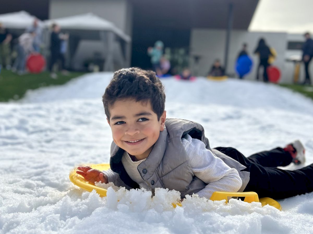 A child is smiling and lying on the snow with a sled, in front of a modern building, with people in the background.