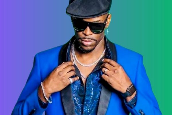 A person in a blue suit and hat, wearing sunglasses and jewelry, poses against a colorful gradient background.