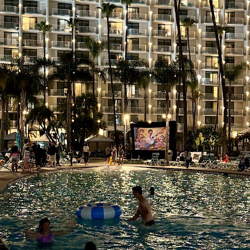 The image shows a night scene at a swimming pool, where people are enjoying the water while a movie is being projected on a screen in the background.