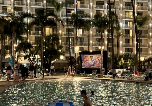 The image shows a night scene at a swimming pool, where people are enjoying the water while a movie is being projected on a screen in the background.